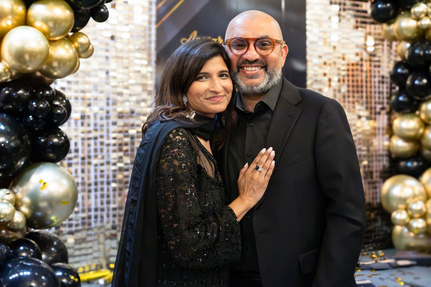 Couple in formal black attire smiling against festive gold balloon backdrop.