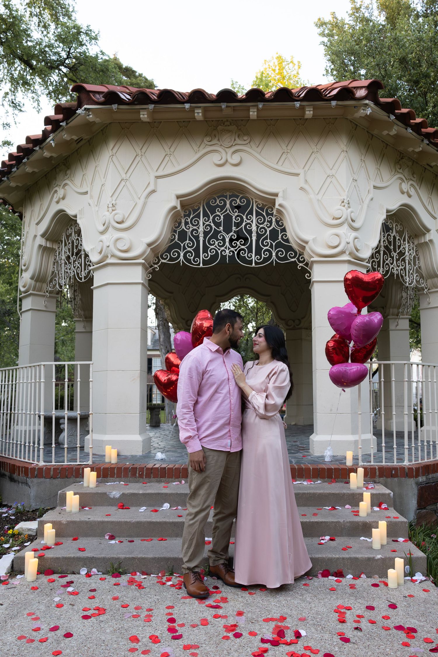 Couple standing before ornate gazebo with heart balloons, rose petals, and candles in a romantic Valentine's Day proposal setting.