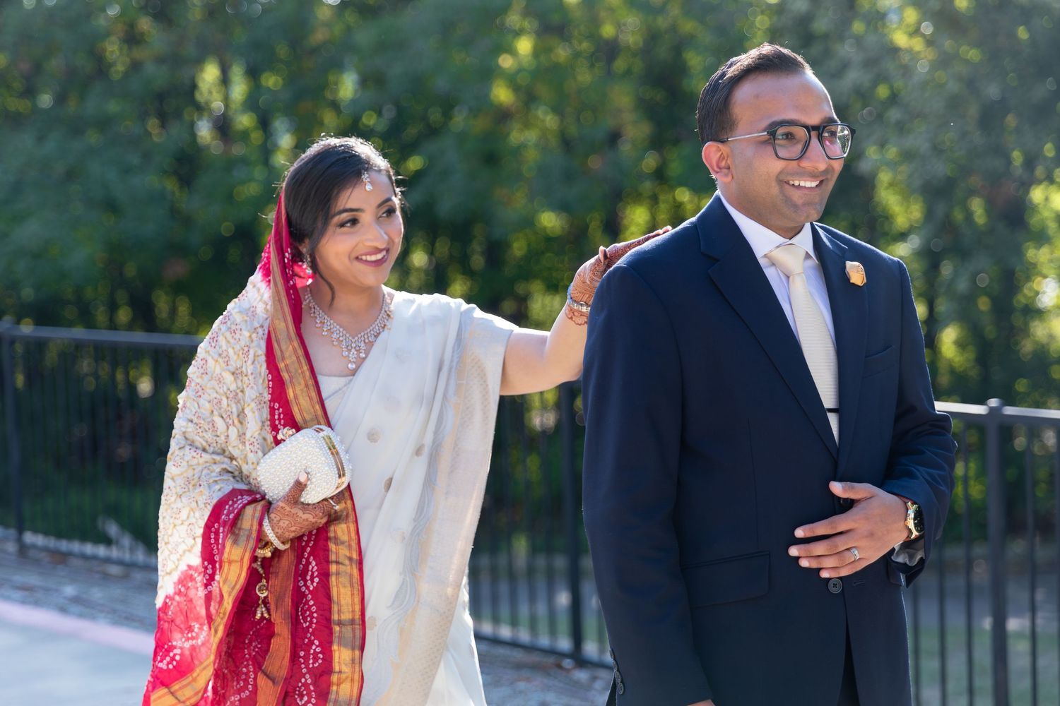 A couple in traditional South Asian wedding attire embraces outdoors during their wedding photoshoot in a garden setting.
