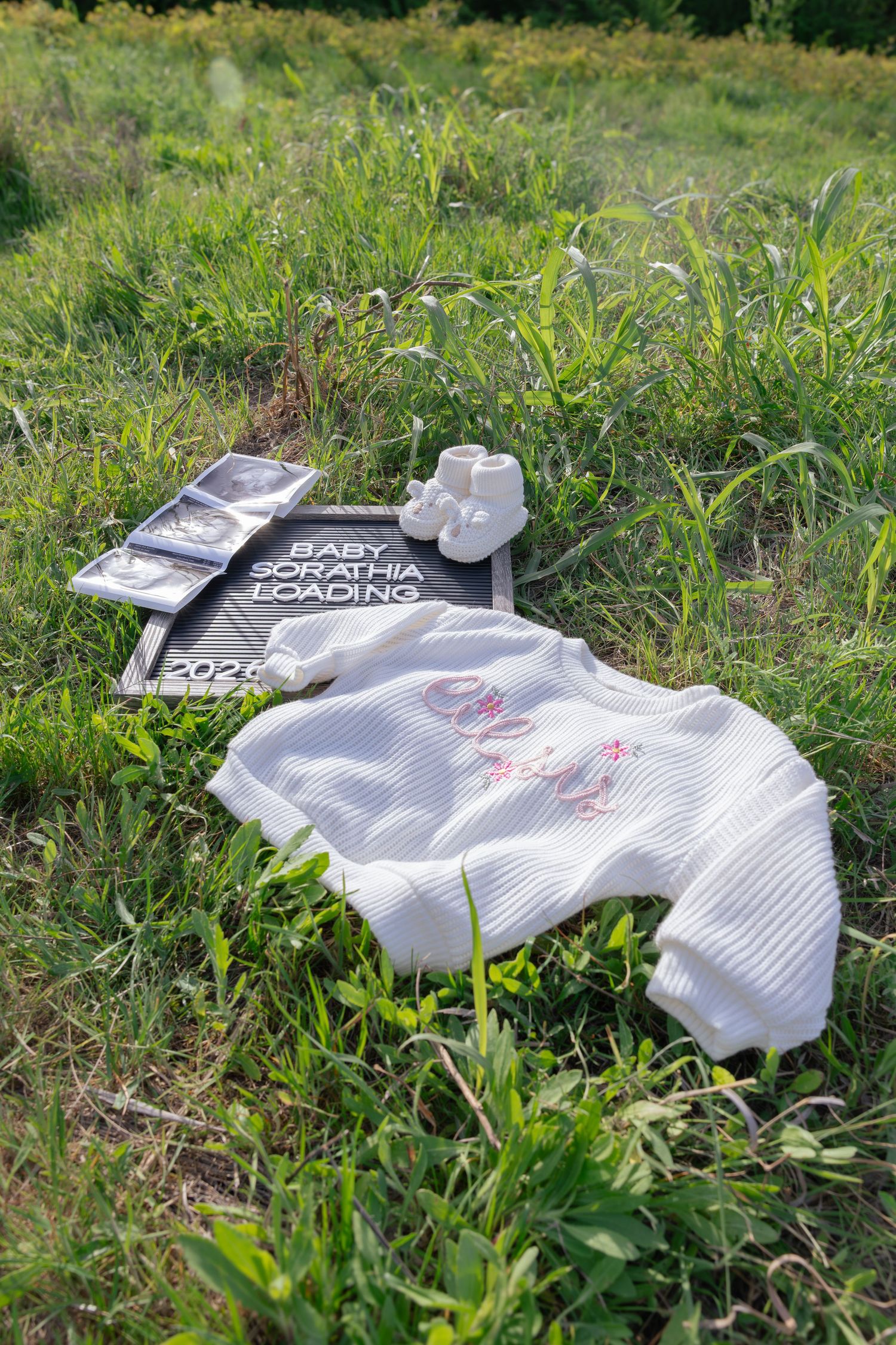Baby announcement flat lay with white knit onesie, booties, ultrasound photos, and a sign reading Baby Sorathia Loading 2024.