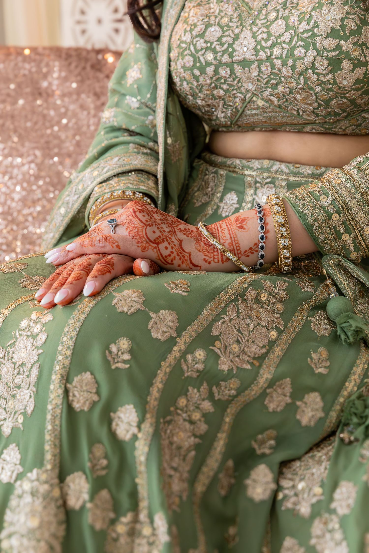Intricate henna design adorns hands against sage green lehenga with ornate silver embroidery and floral patterns.