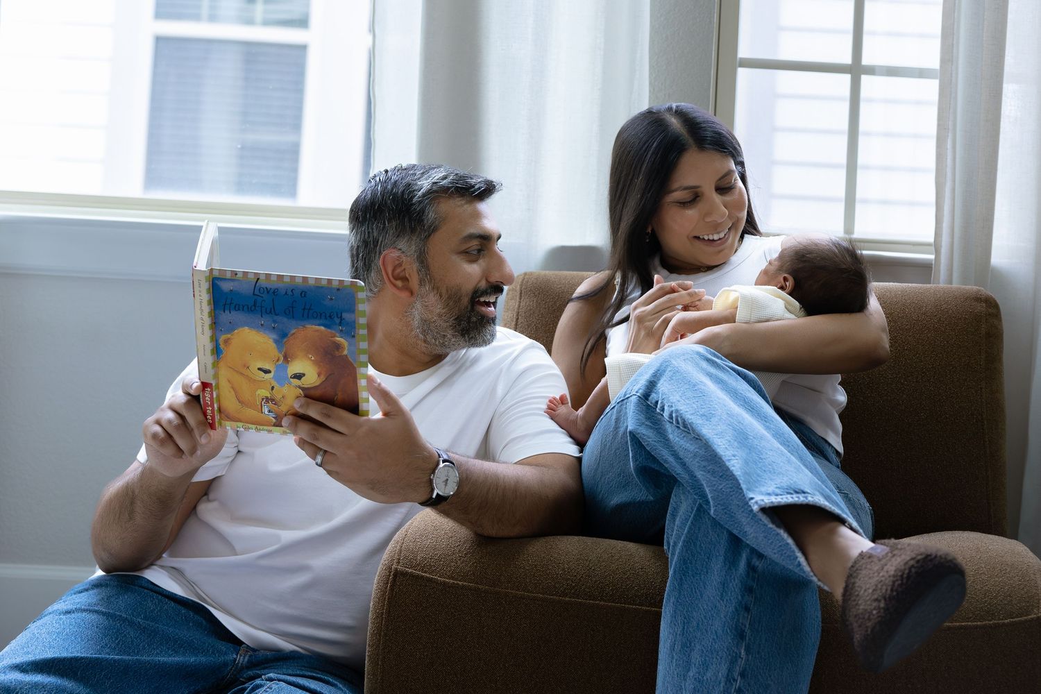 A man reads a book while a woman breastfeeds a newborn baby, sitting together on a cozy armchair at home.