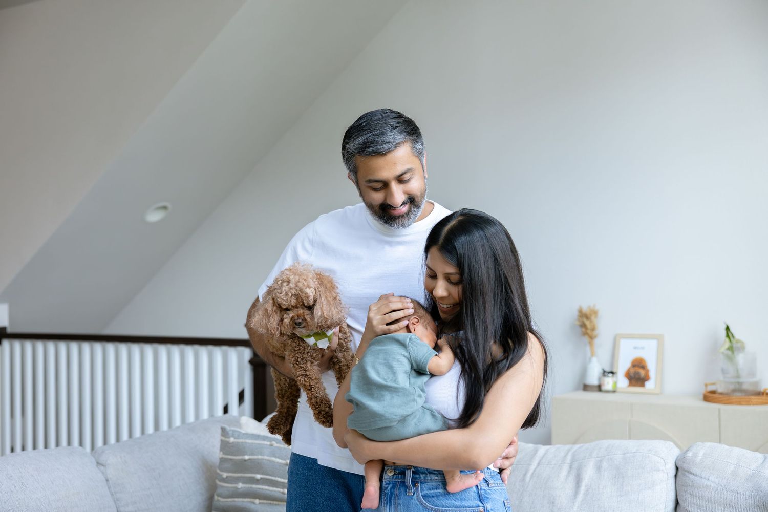 A happy family with a man, woman, and baby cuddle together indoors while holding an adorable brown poodle dog.