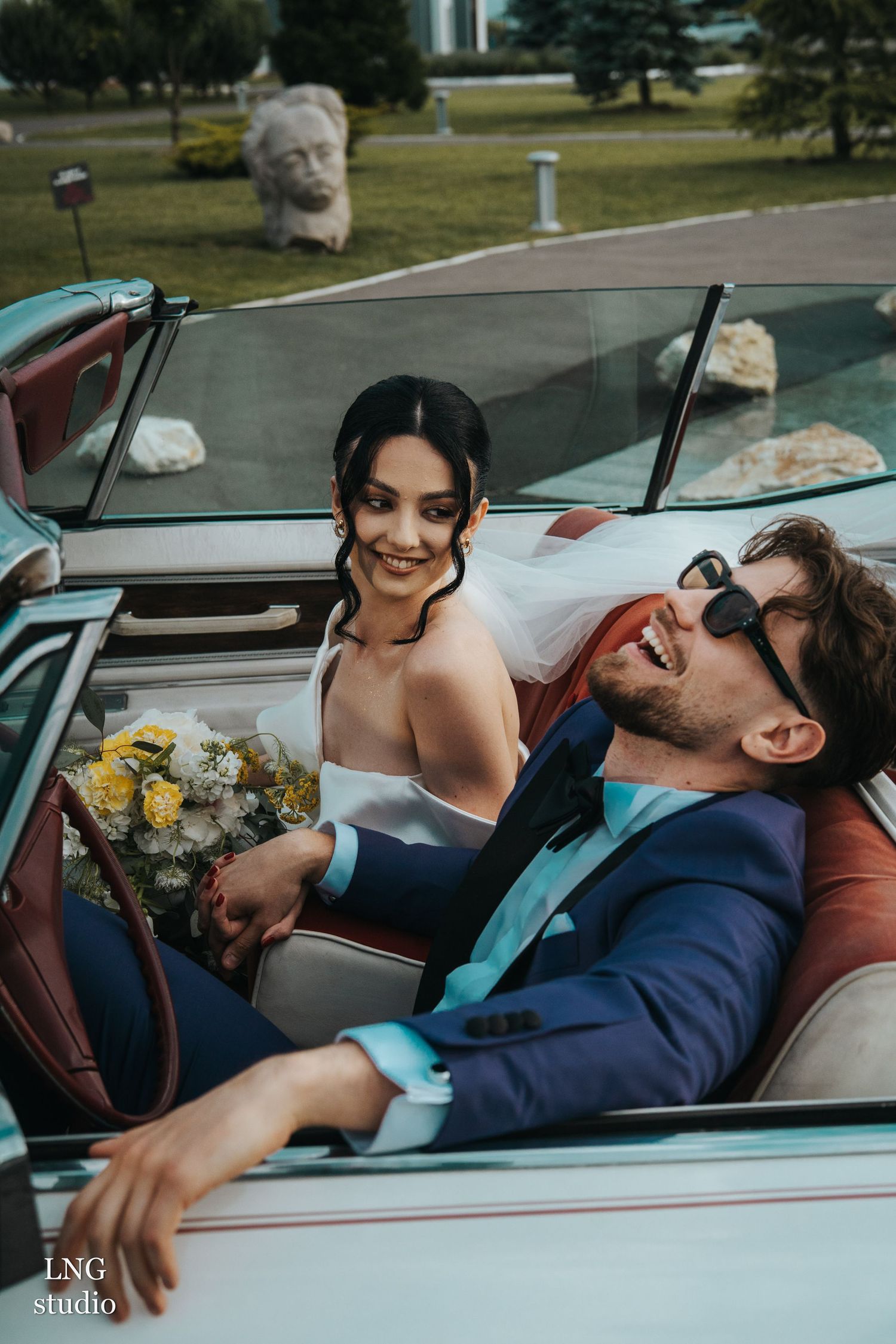 A joyful wedding couple sharing a candid moment in a vintage convertible car with yellow flowers.
