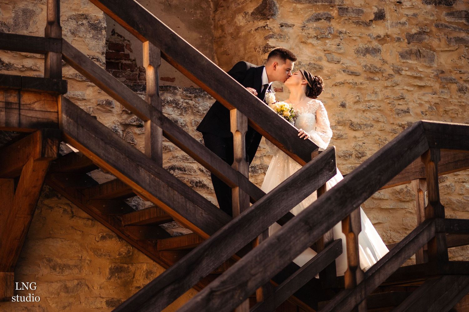 Wedding couple poses on rustic wooden staircase against stone wall backdrop.