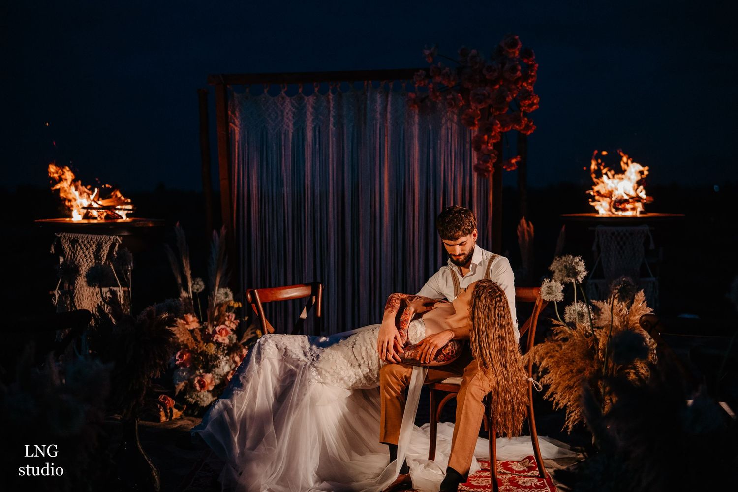 A couple sits together at a stylishly decorated outdoor wedding setup with fire bowls and floral arrangements at night.