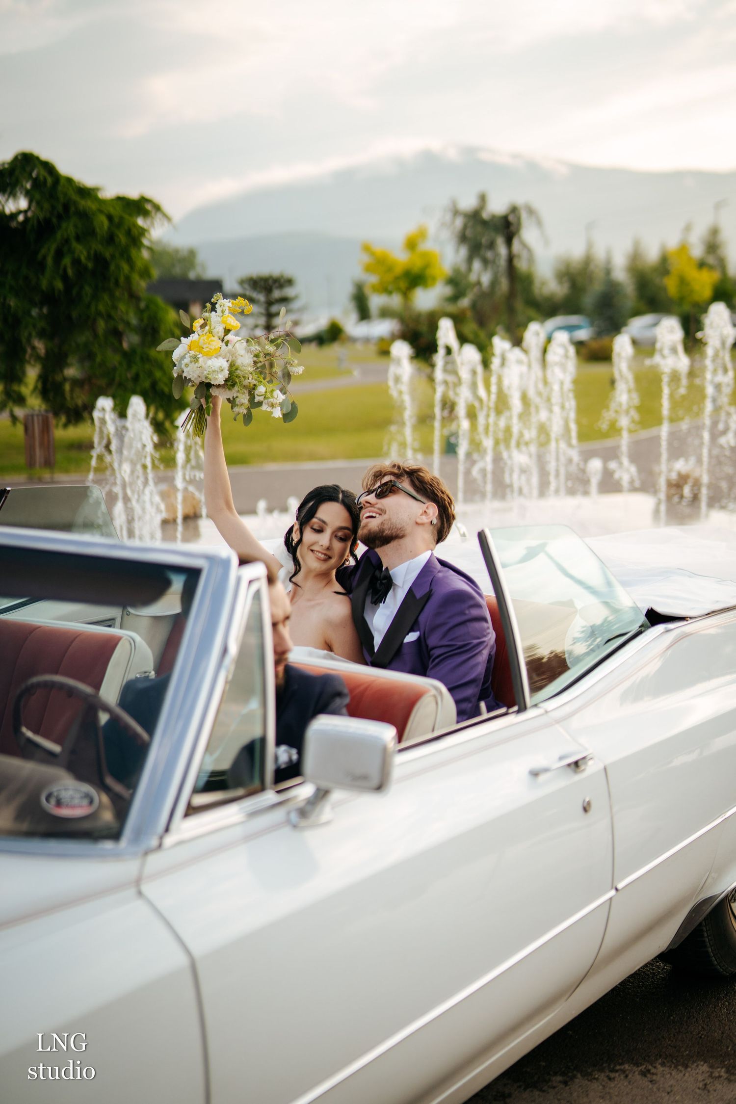 Romantic newlyweds pose in a white vintage convertible with fountains and mountains in the background.