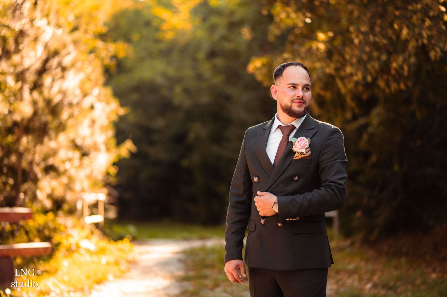Groom in formal black suit with boutonniere poses in autumn setting with golden foliage.