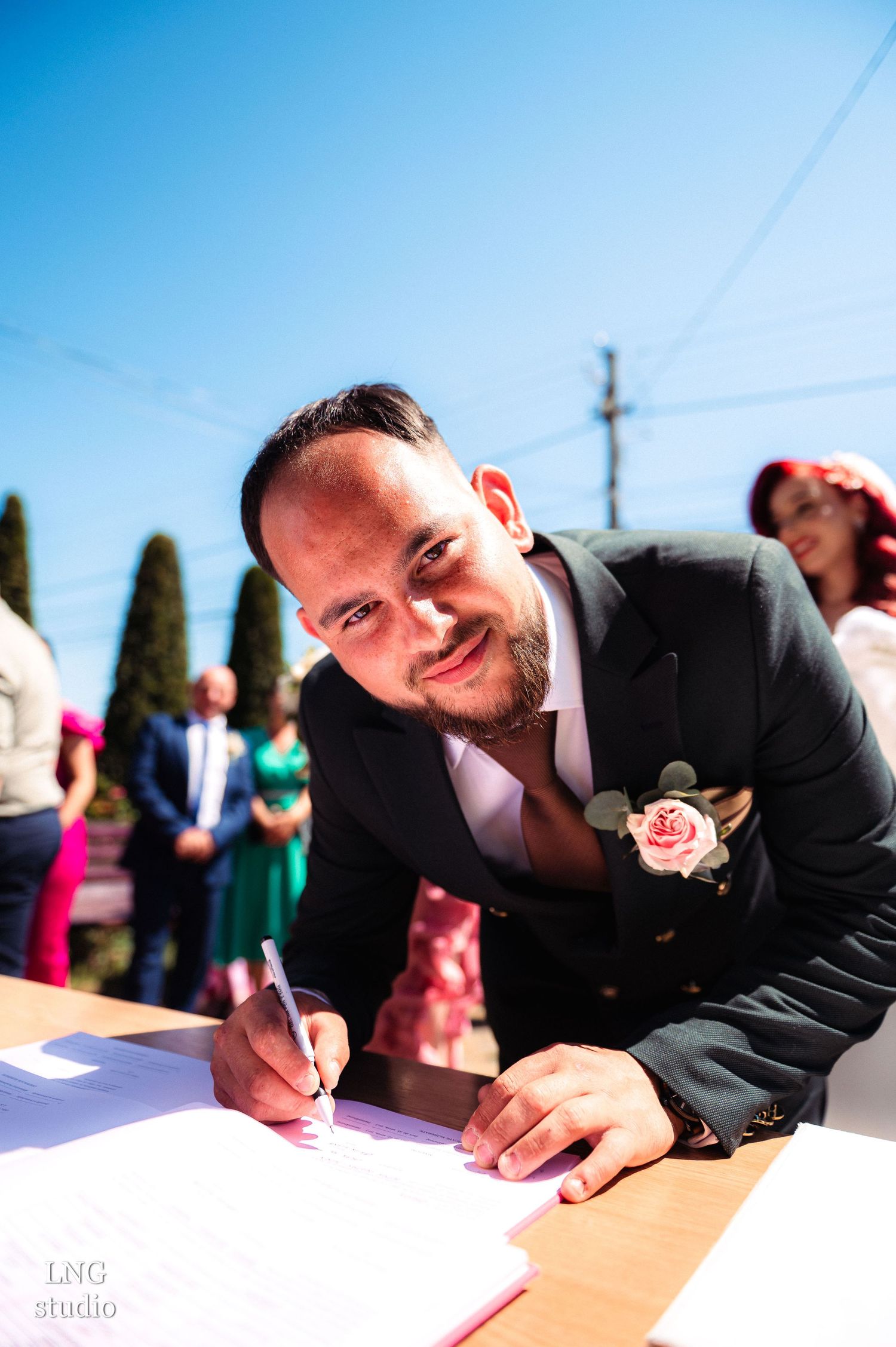 Groom signs wedding documents at outdoor ceremony table under bright blue sky.