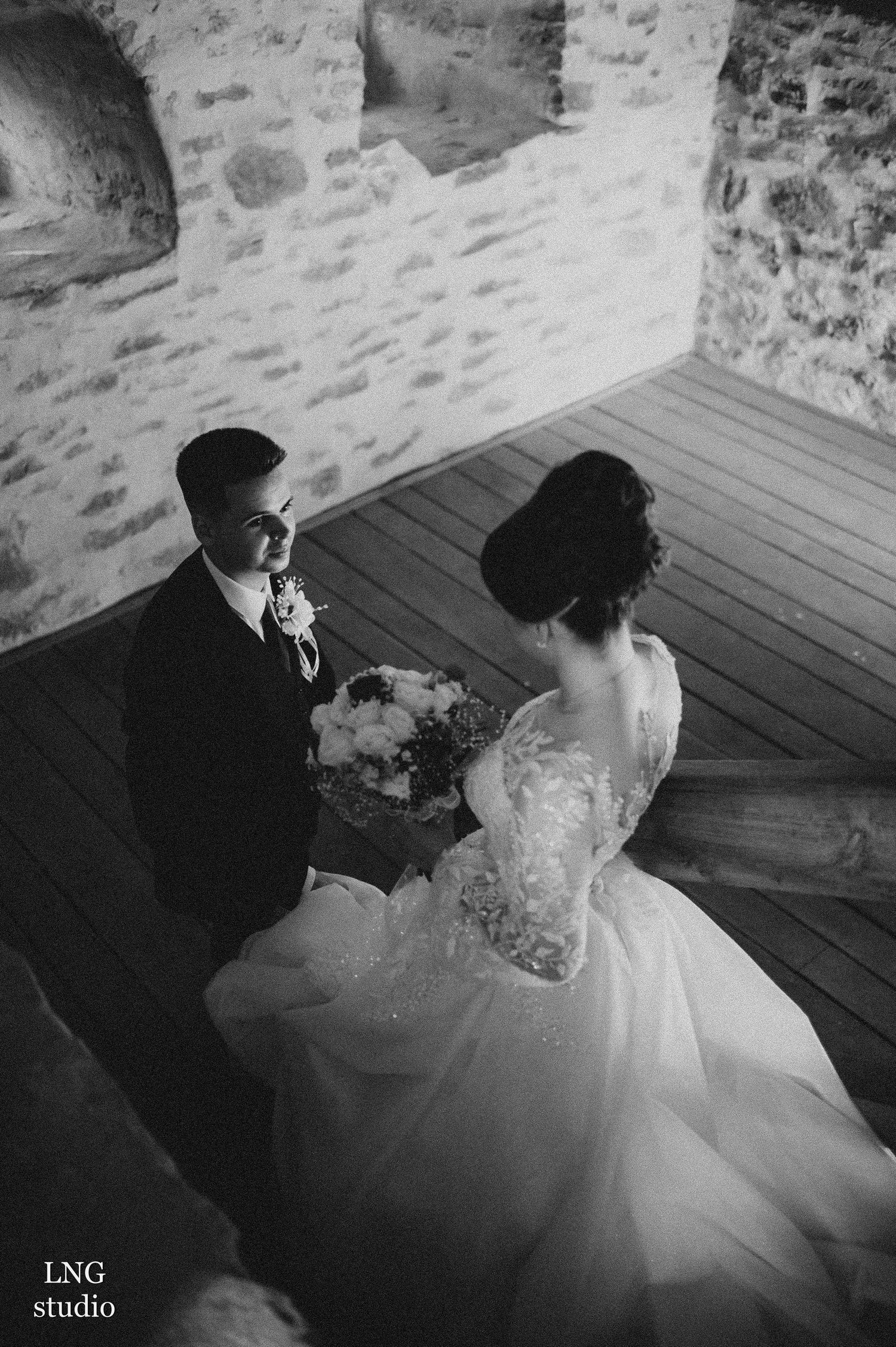 Black and white photo of a bride in lace gown with groom sitting on wooden floor in rustic stone venue.