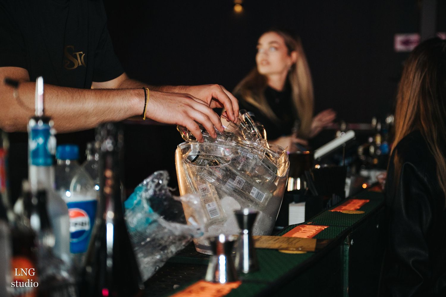 Dimly lit bar scene showing hands preparing drinks with various bottles and glasses.