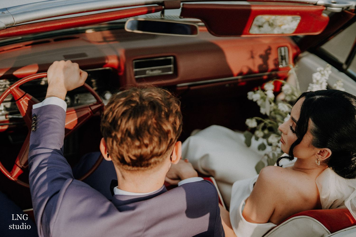 Wedding couple shares intimate moments inside a vintage red car decorated with floral arrangements.
