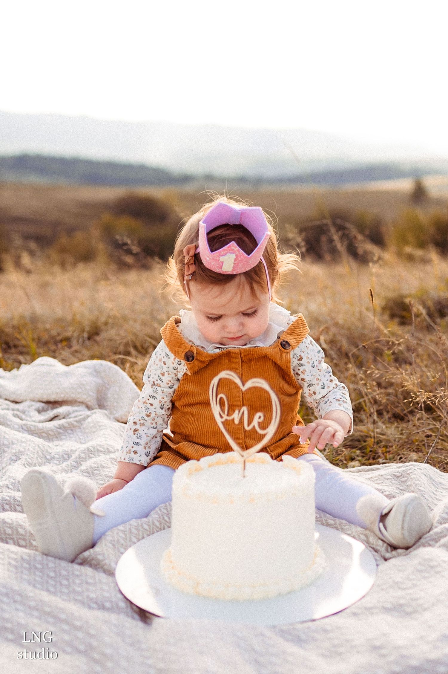 Toddler in orange dress looks down at white birthday cake with heart decoration.