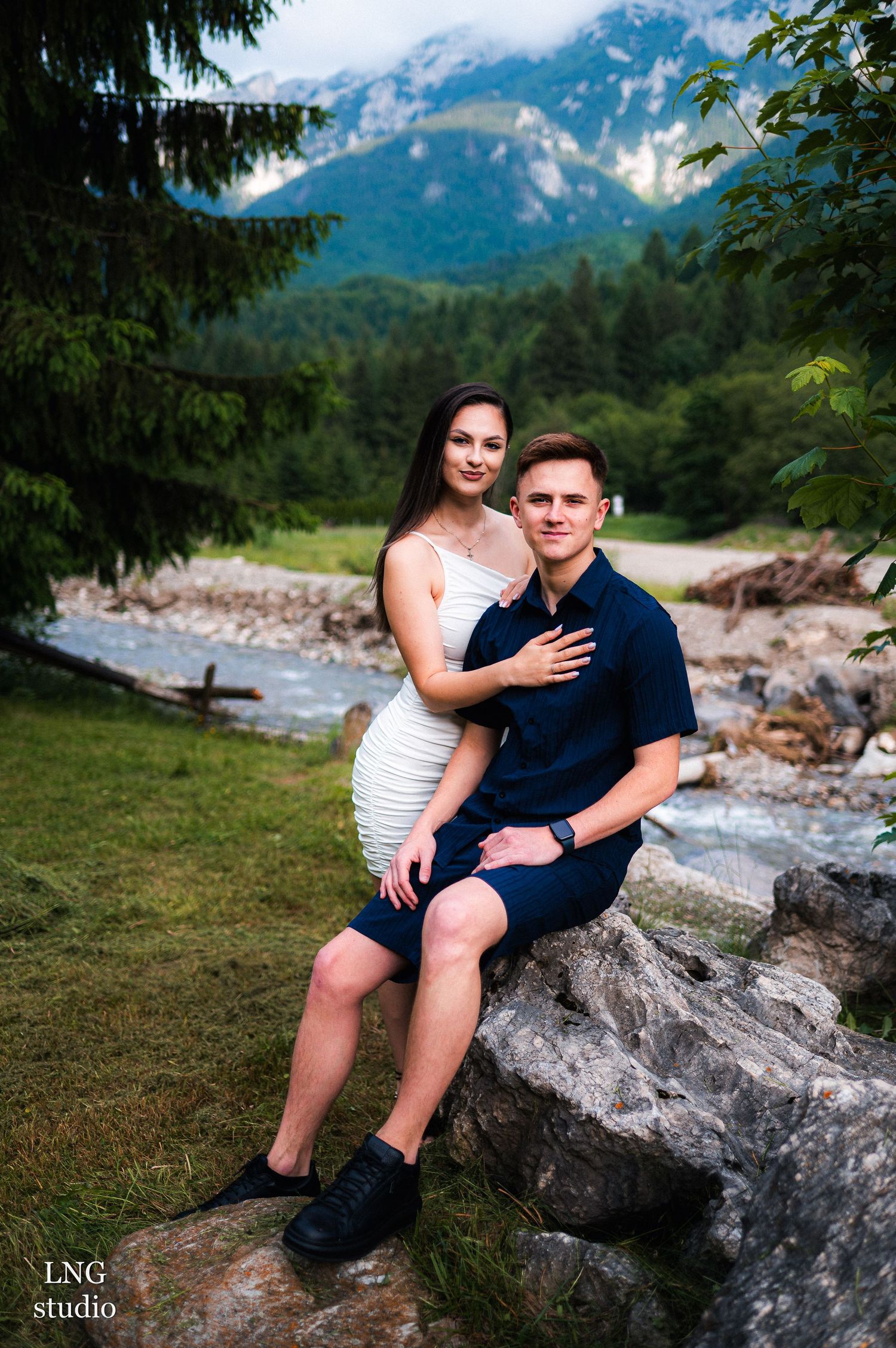 A couple poses on rocky terrain with stunning mountain views and a stream in the background.