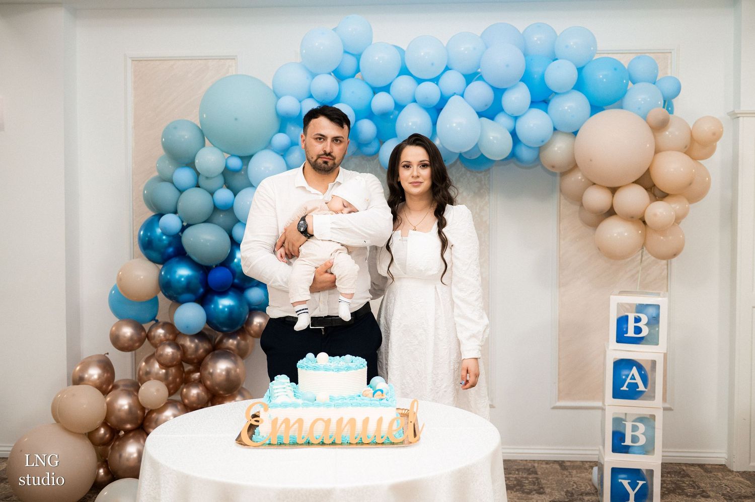 A celebration setup with blue and beige balloon arch, cake on white table, and decorative baby blocks.