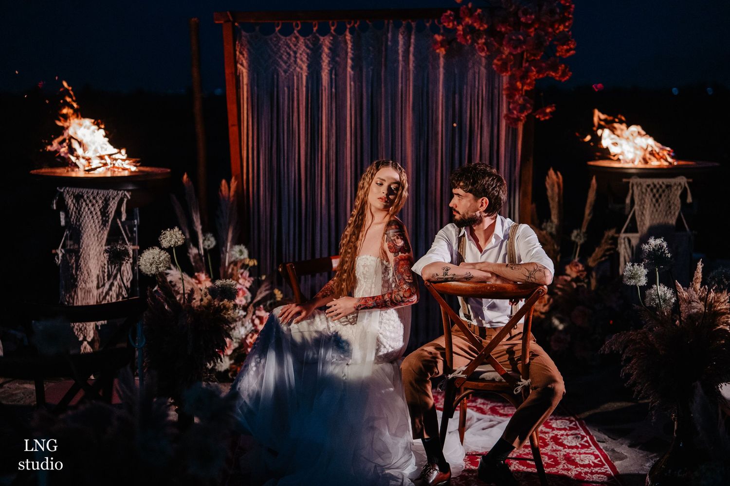 A moody nighttime wedding scene with flaming torches, draped fabric backdrop, floral decor and boho styling.