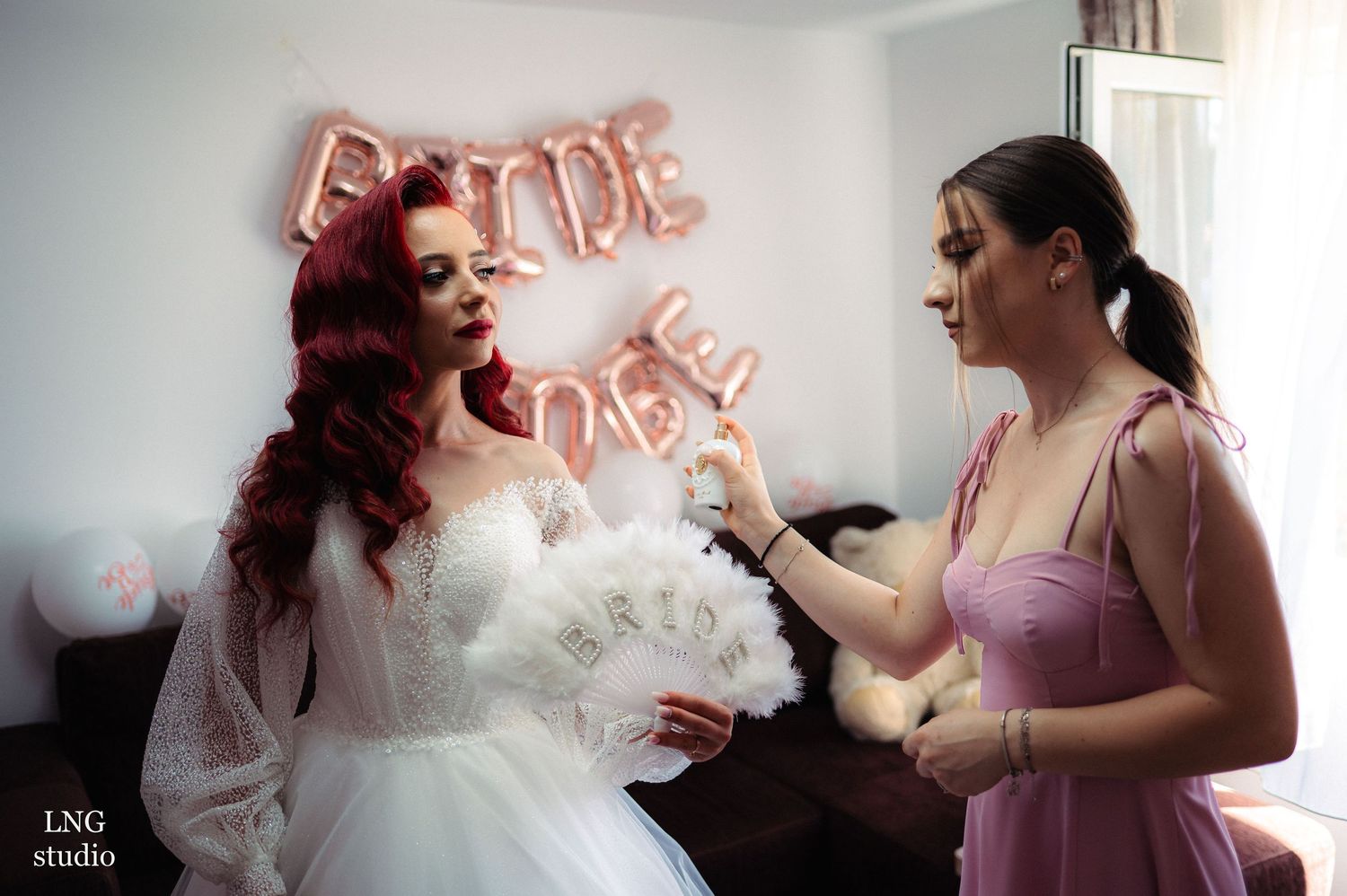 Pink-robed bridesmaids help prepare for wedding celebrations against white decor and rose gold balloon lettering.