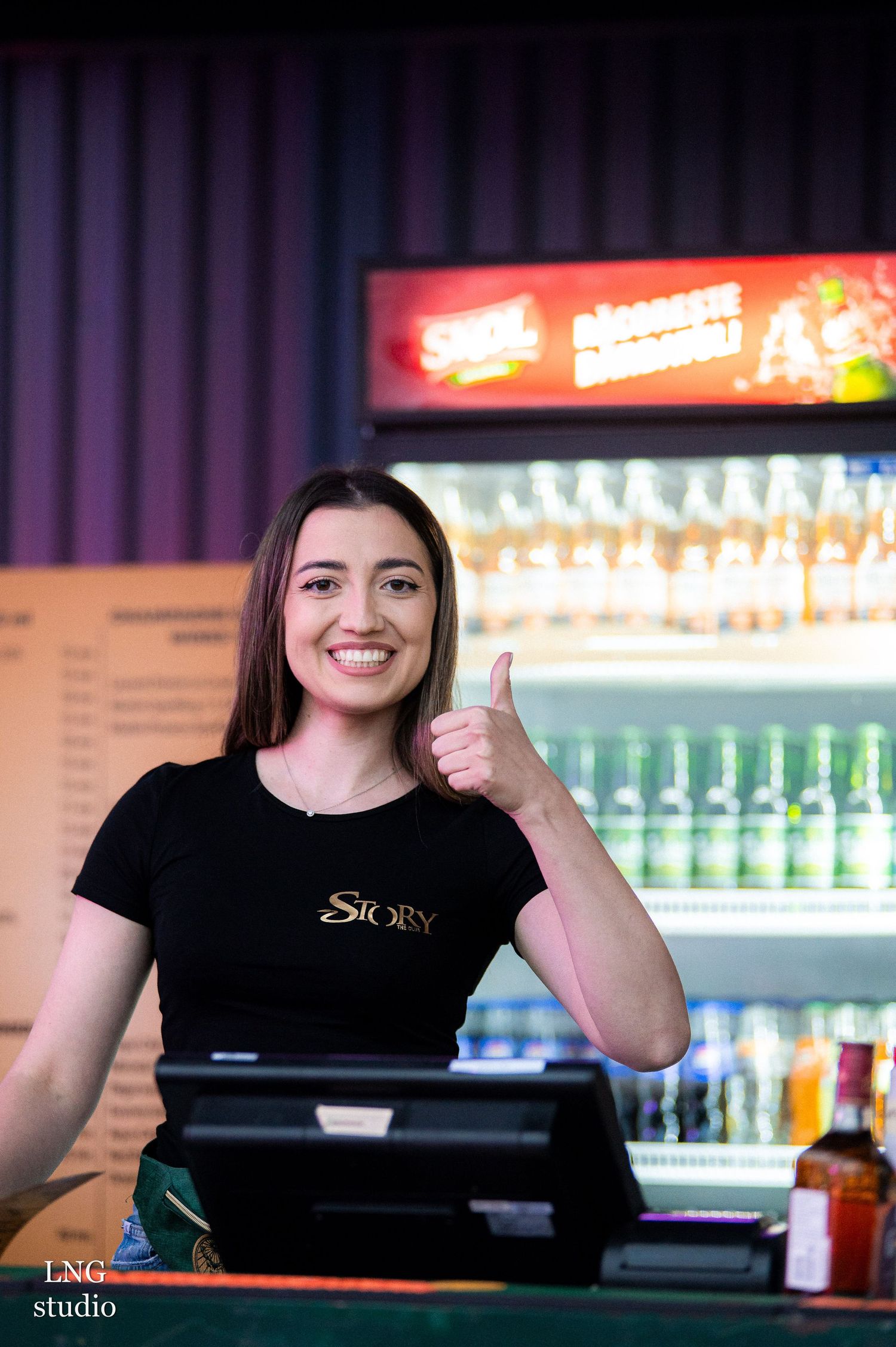 A friendly bartender wearing a black t-shirt gives thumbs up while working behind the counter with beverages displayed.