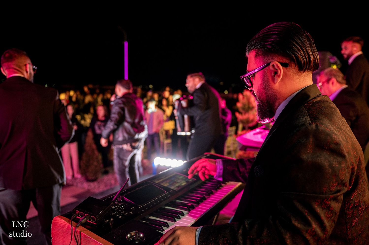 Musician plays keyboard at nighttime concert with colorful stage lighting and crowd in background.