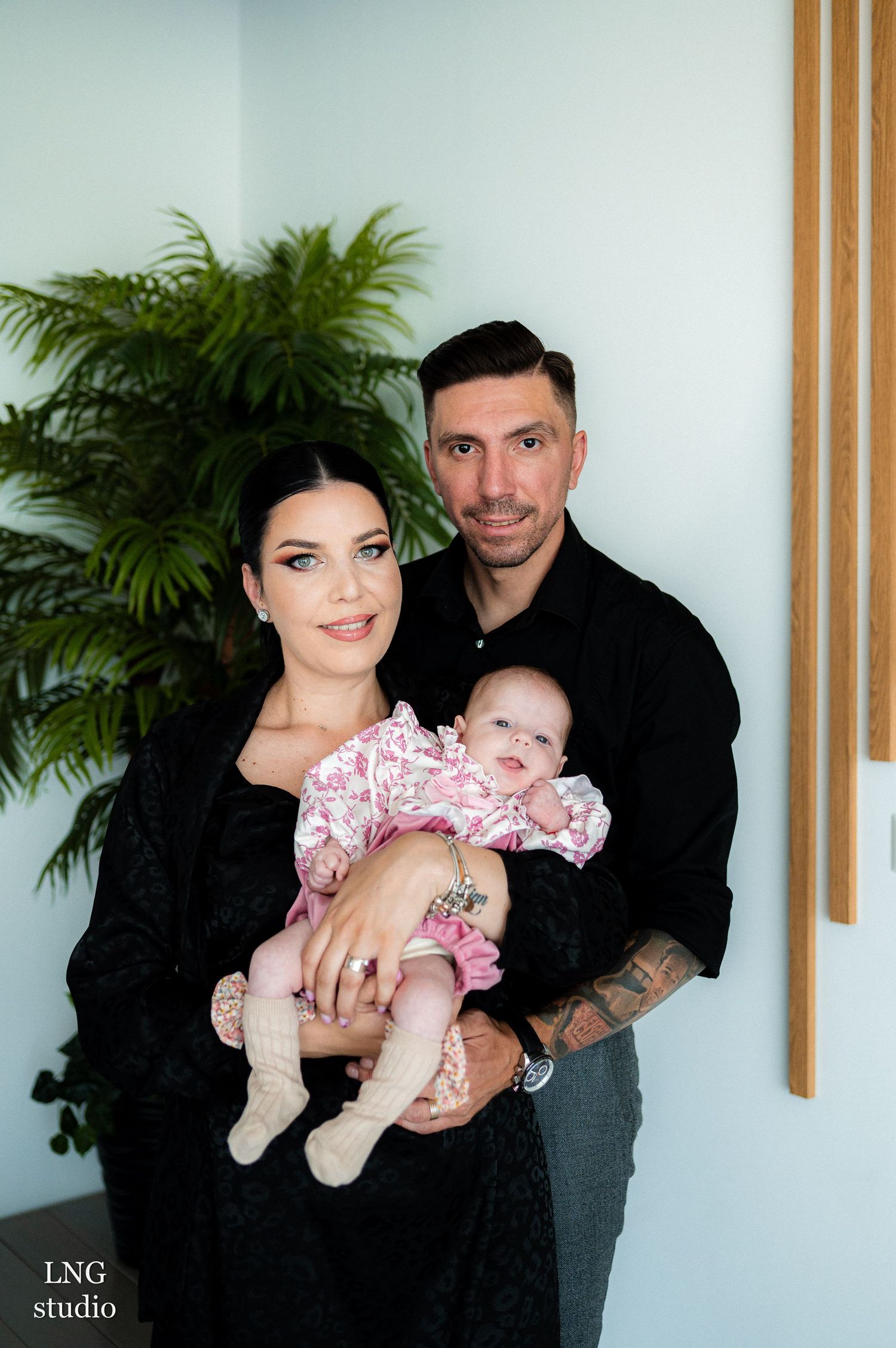 Parents cradle infant wearing pink ruffled outfit against light blue wall with wood accents.