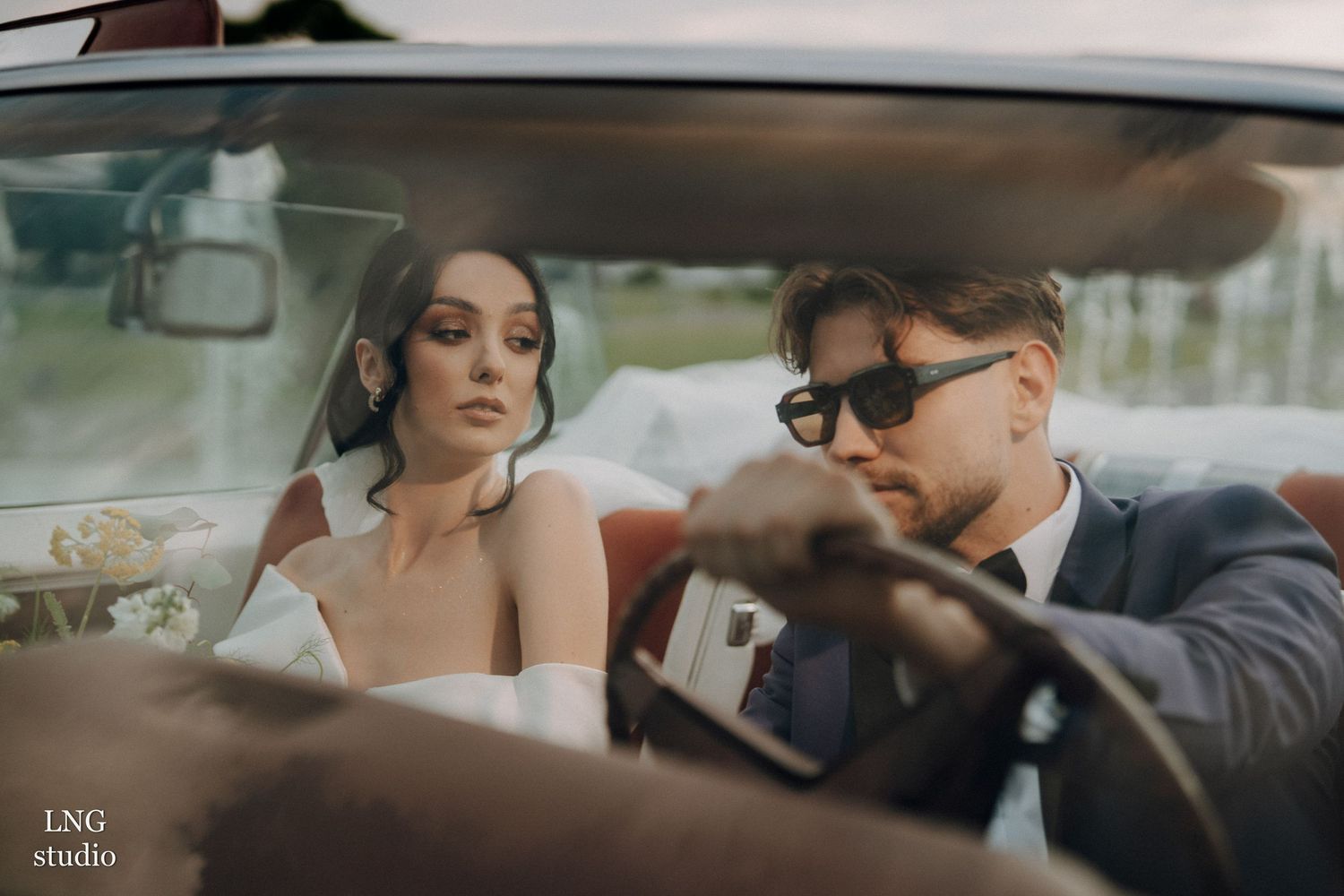 A couple sits in a vintage car, with the driver in a suit and sunglasses while his passenger wears bridal attire.