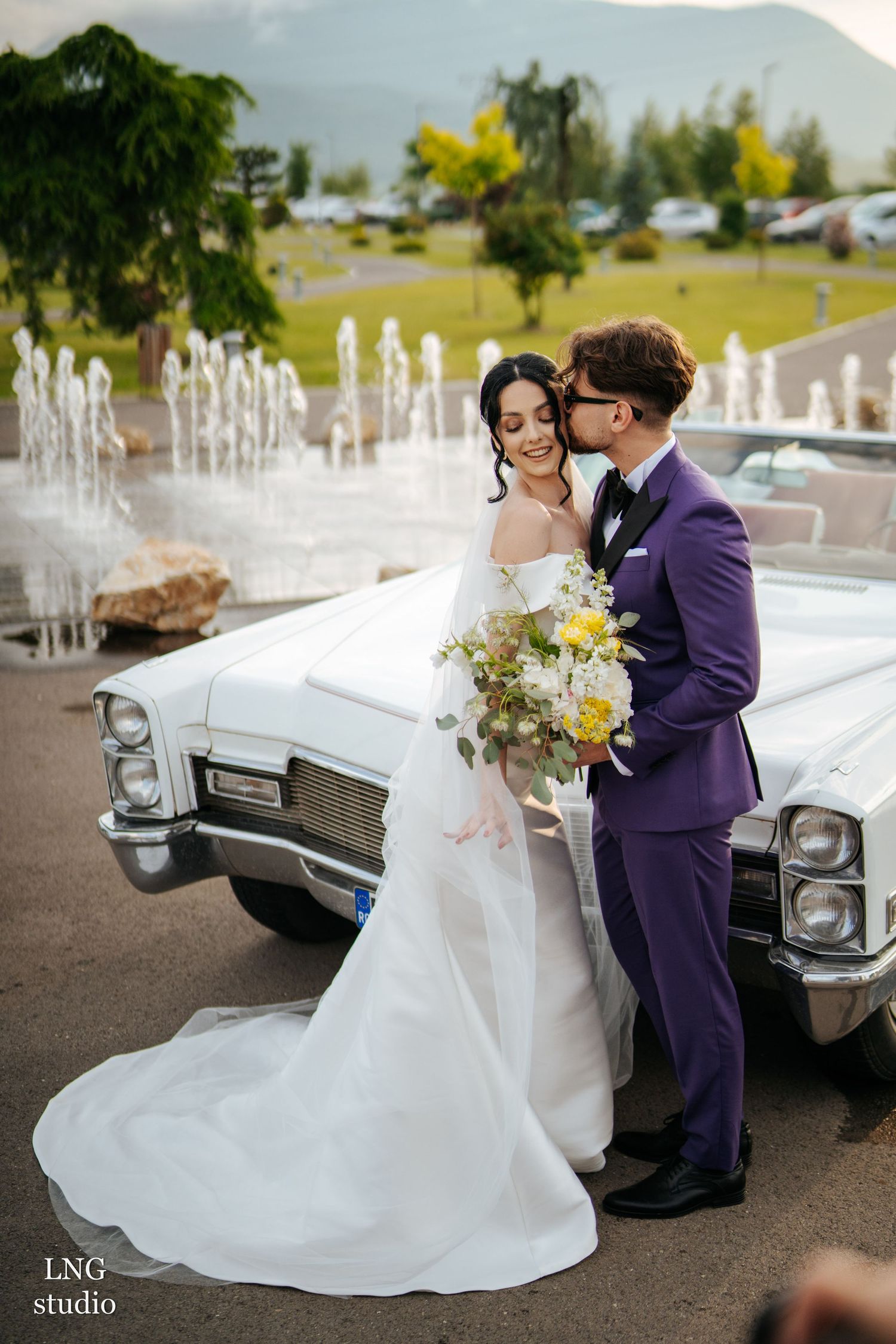 Wedding couple poses near a vintage car with water fountains and mountains in the backdrop.