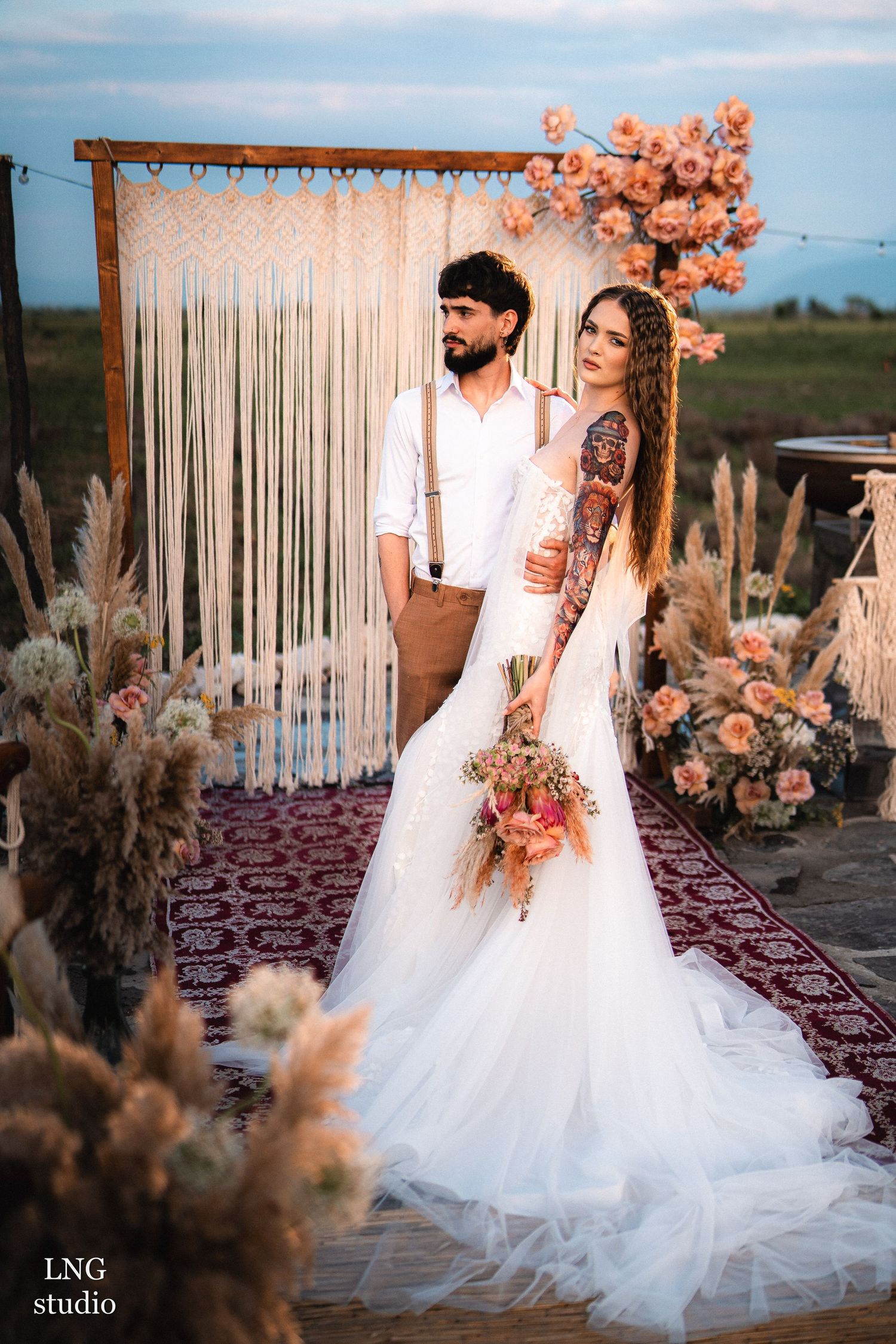 Elegant outdoor wedding photoshoot at sunset with floral archway incorporating pampas grass and coral blooms on Persian rug.