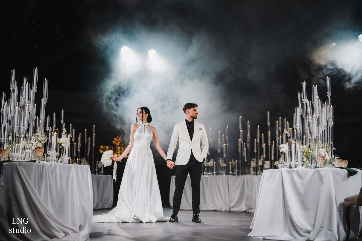 A wedding couple poses under sparkling crystal chandeliers during their elegant reception at a luxurious venue.