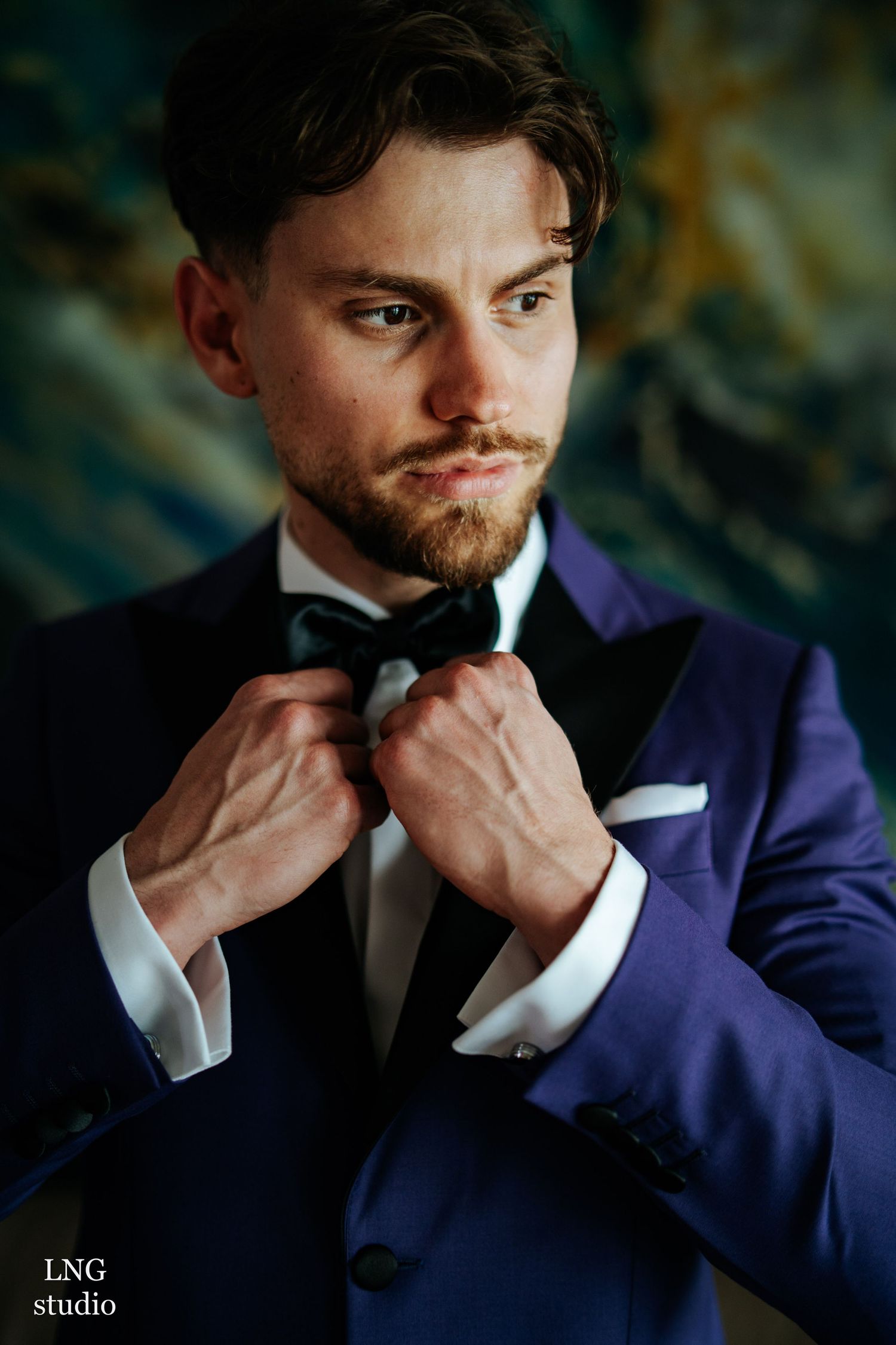 Fashionable portrait of a gentleman in a dark blue tuxedo adjusting his bowtie against a blurred background.