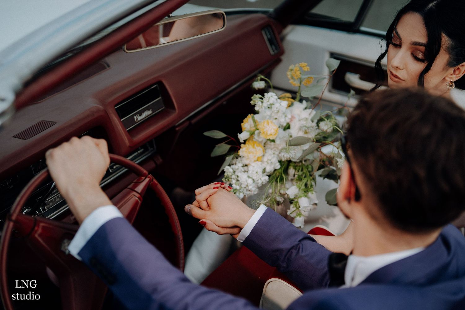 Wedding party members share happy laughter while seated in a vintage car's interior with floral bouquets.