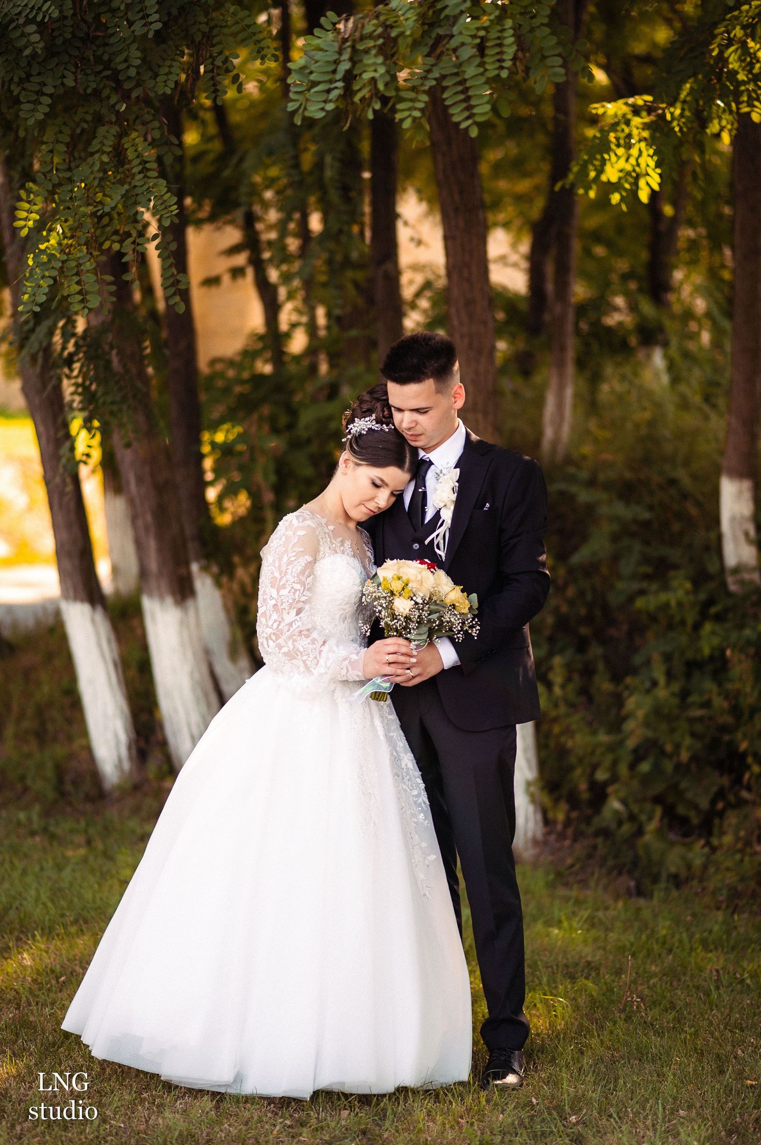 Wedding couple poses among trees with bride holding yellow and white bouquet.