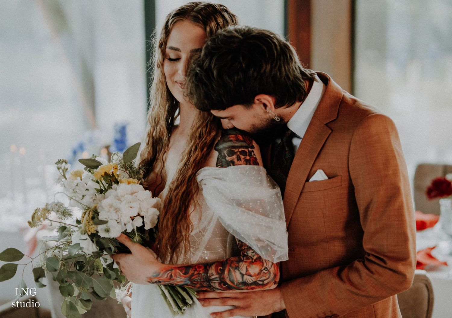 Couple in an autumn wedding setting with bride in white and groom in burnt orange suit share an intimate moment.