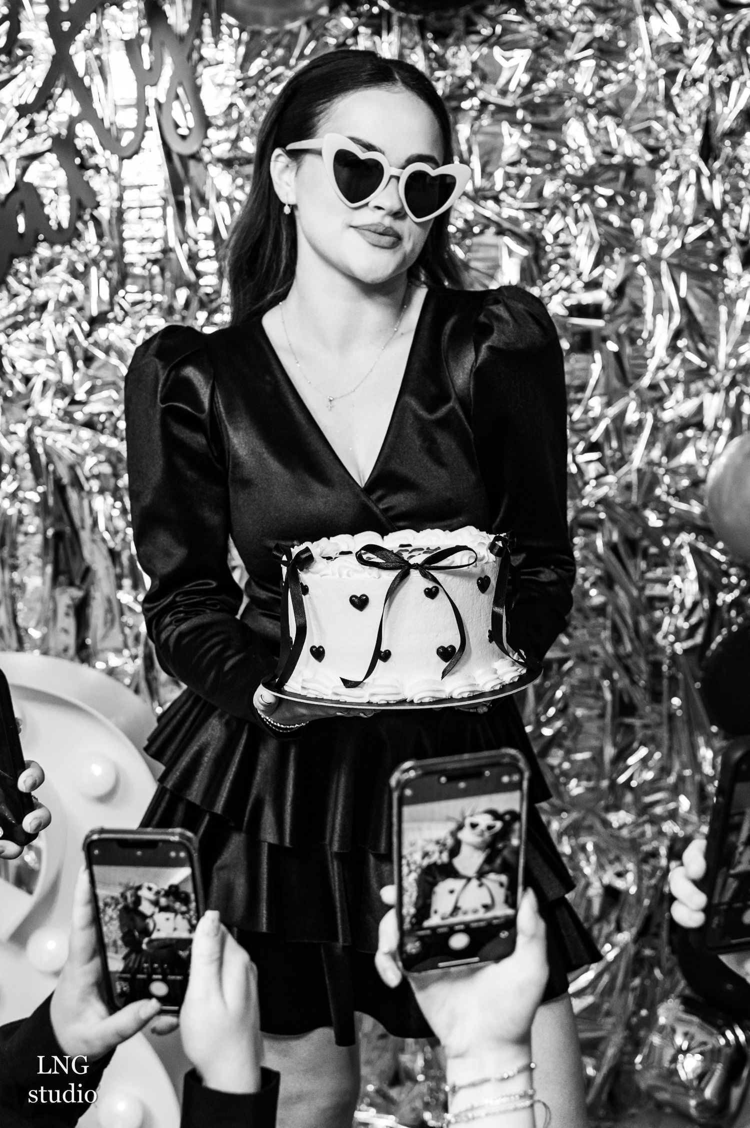 A fashionable woman in black dress and heart sunglasses holds a cake while phones capture photos in black and white.
