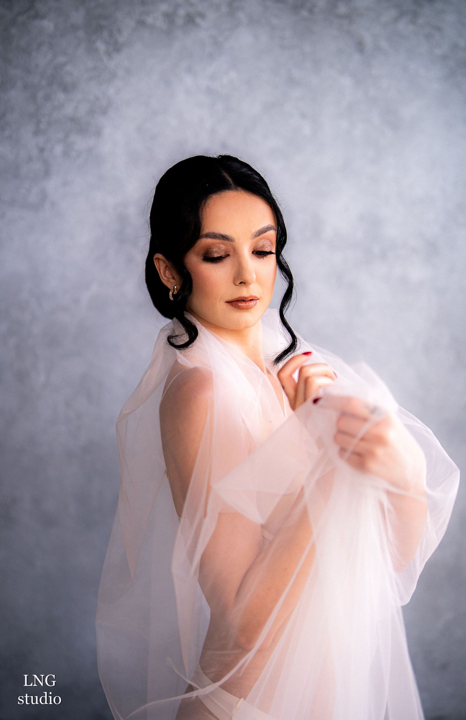 A woman in soft white fabric poses gracefully against a misty gray backdrop in an ethereal bridal portrait series.