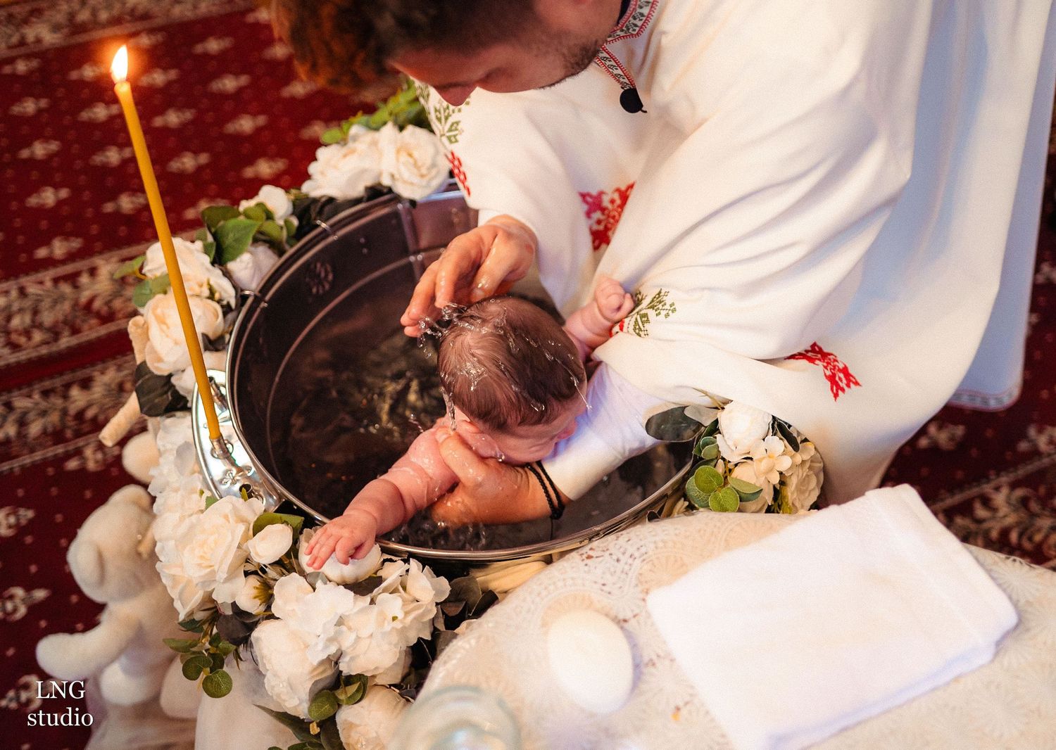 A religious baptism ceremony with candles and white flowers surrounding a baptismal font.