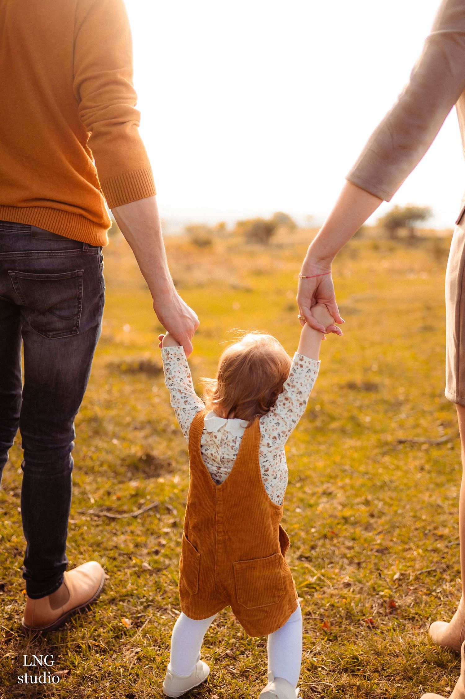 A small child in a brown corduroy dress holds hands with adults while walking in a golden sunlit field.