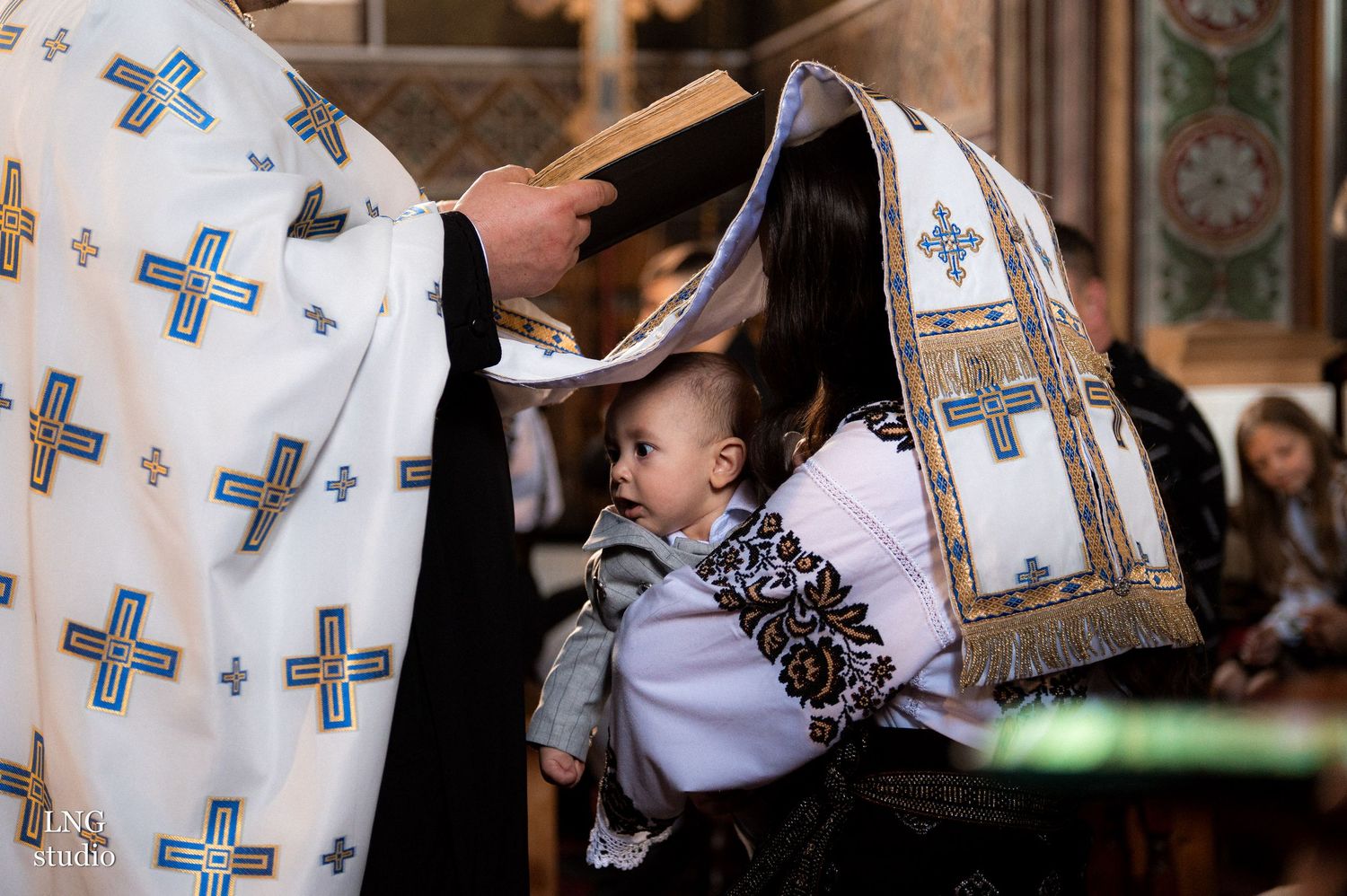 Orthodox priest performing baptism ritual with baby under ceremonial cloth.