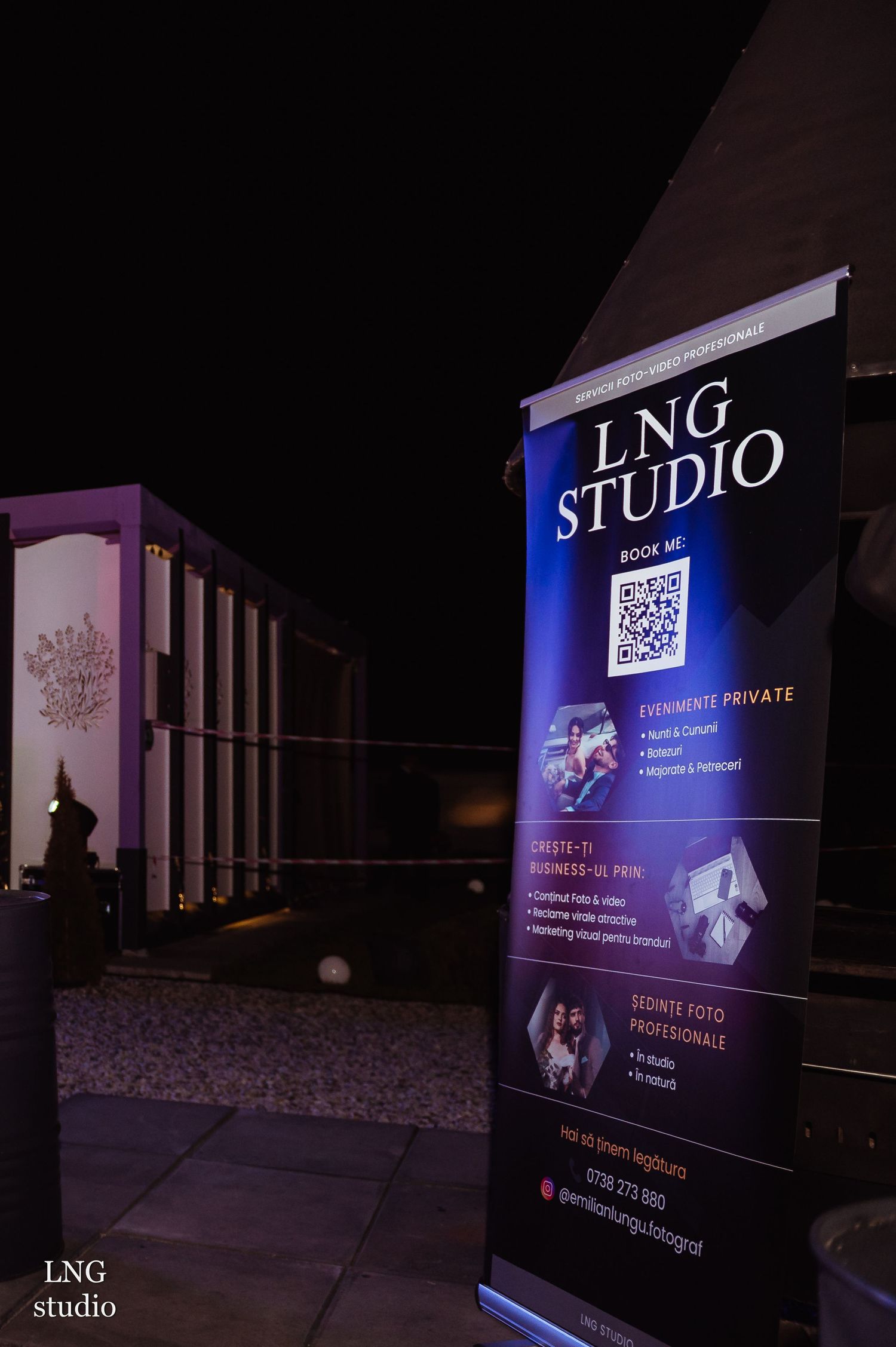 A blue and purple illuminated banner stand for LNG Studio displays business services and QR code outside at night.