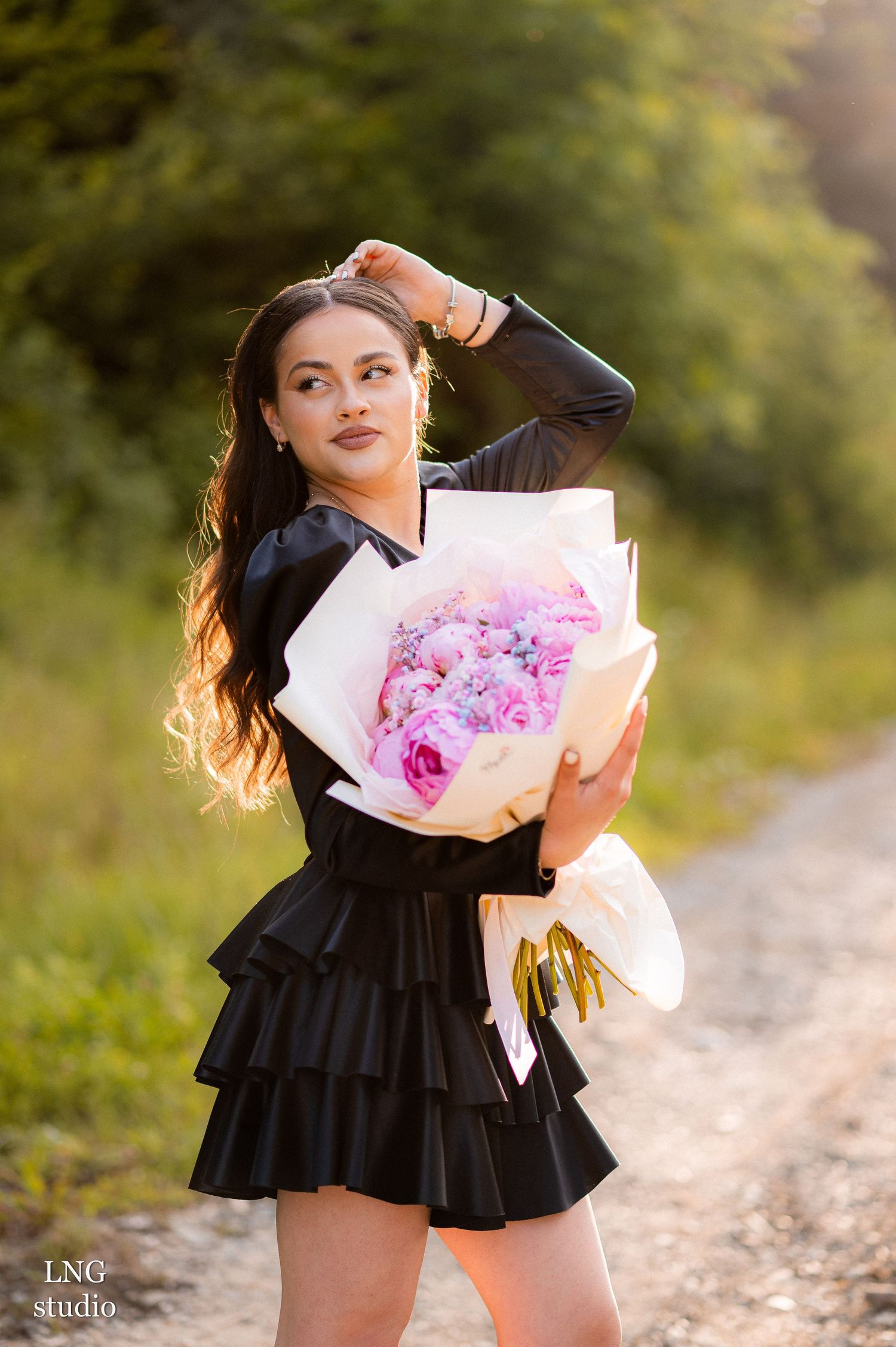 Woman in black dress clutching a pink bouquet of flowers in an outdoor nature setting with soft bokeh background effect.