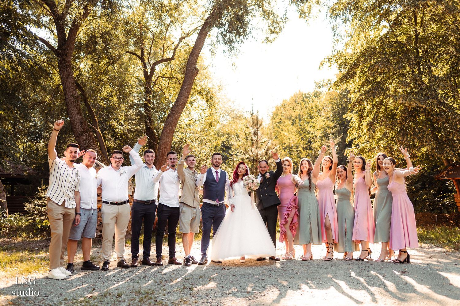 Wedding party and guests celebrate with arms raised outdoors under tall trees.