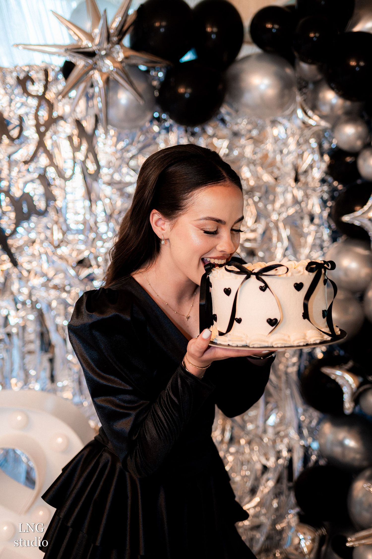 A joyful celebration scene with a black evening dress against silver and black balloon decorations and a decorated cake.