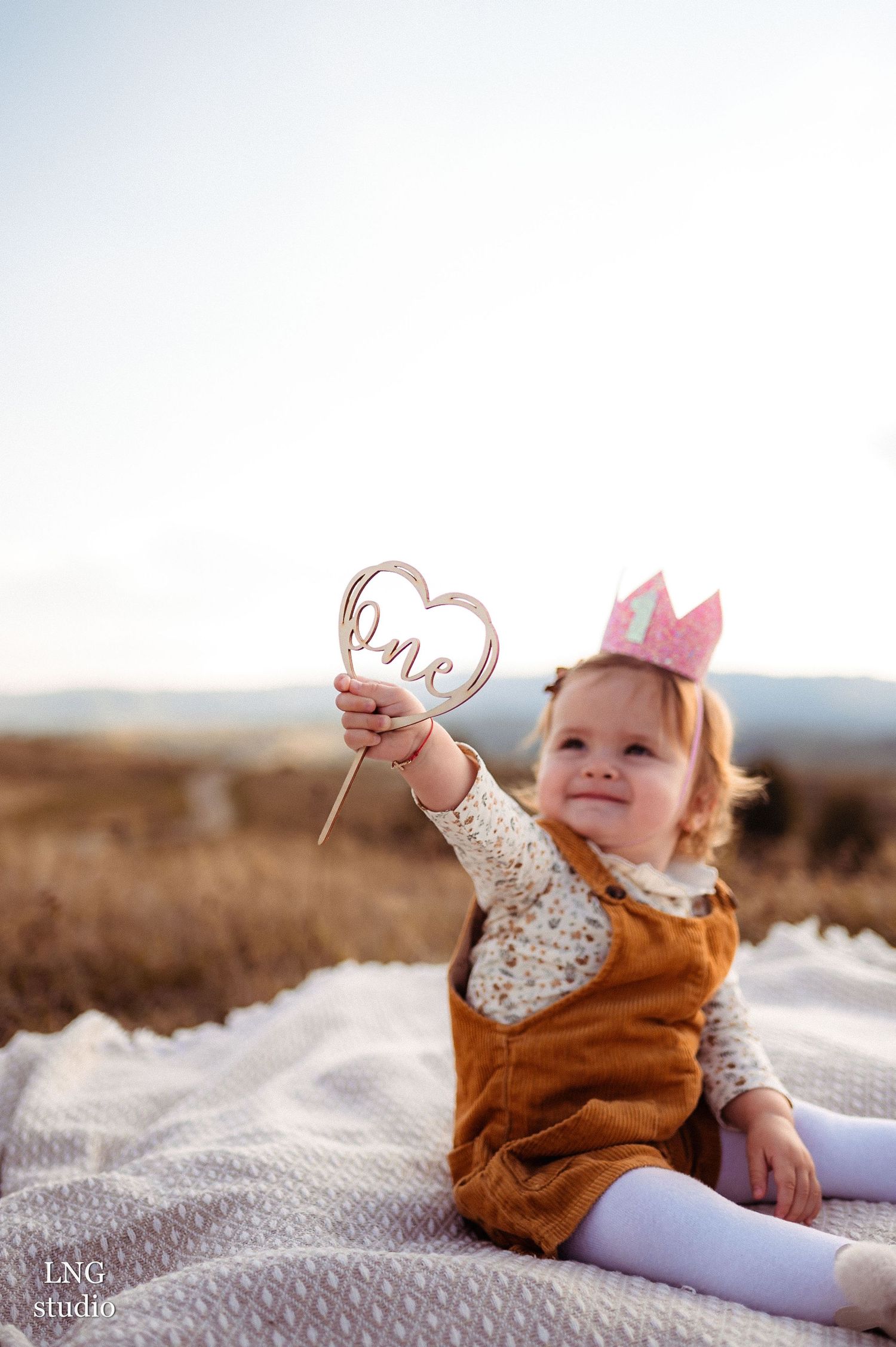 Toddler in brown overalls holding heart wand while sitting on blanket outdoors.