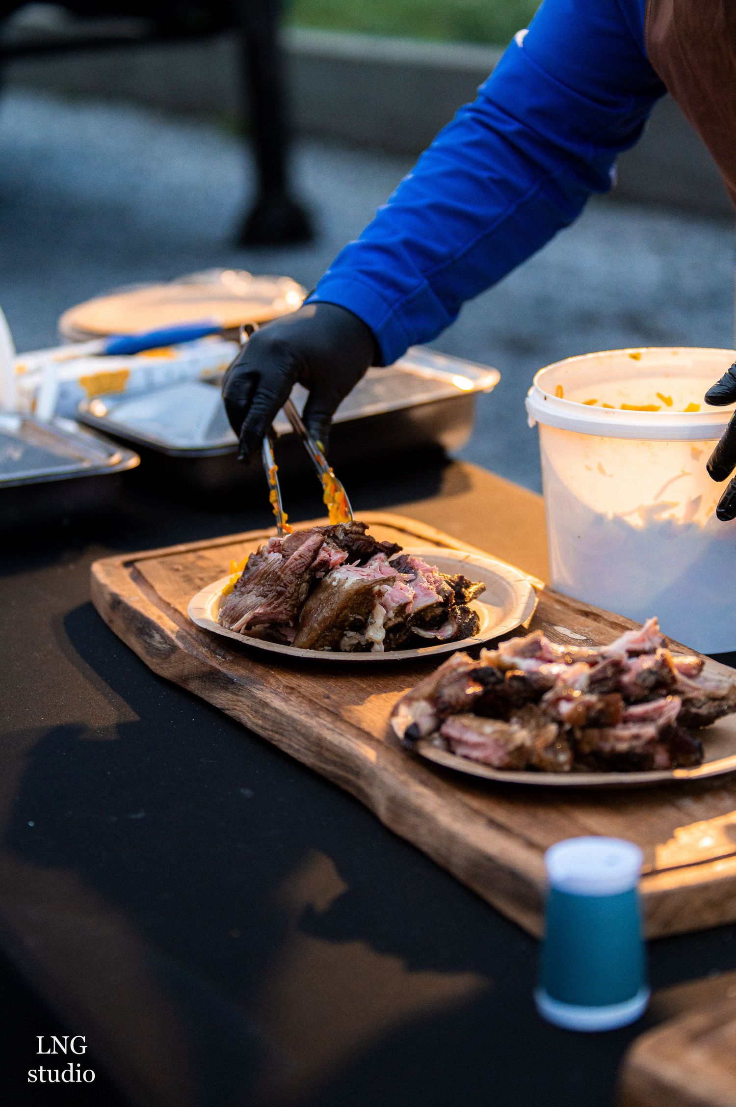 Person in blue sleeve and black gloves plates grilled meat on wooden serving boards at outdoor event.