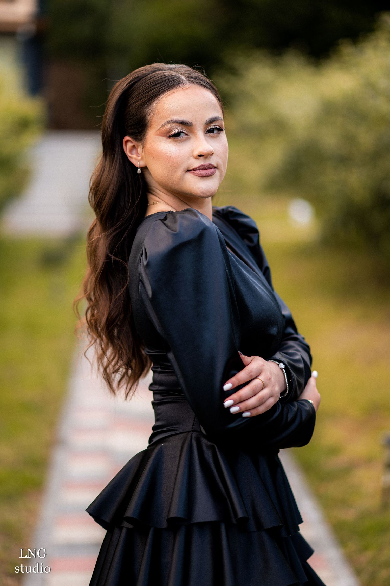A woman in an elegant black dress with long wavy dark hair poses outdoors against a blurred natural background.
