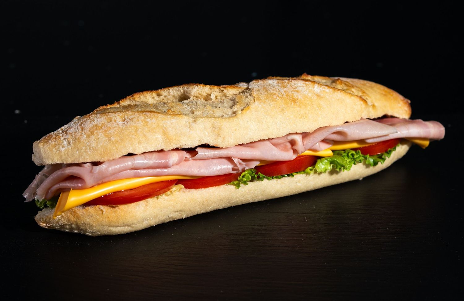 Long crusty baguette sandwich filled with fresh ingredients shown on dark background.