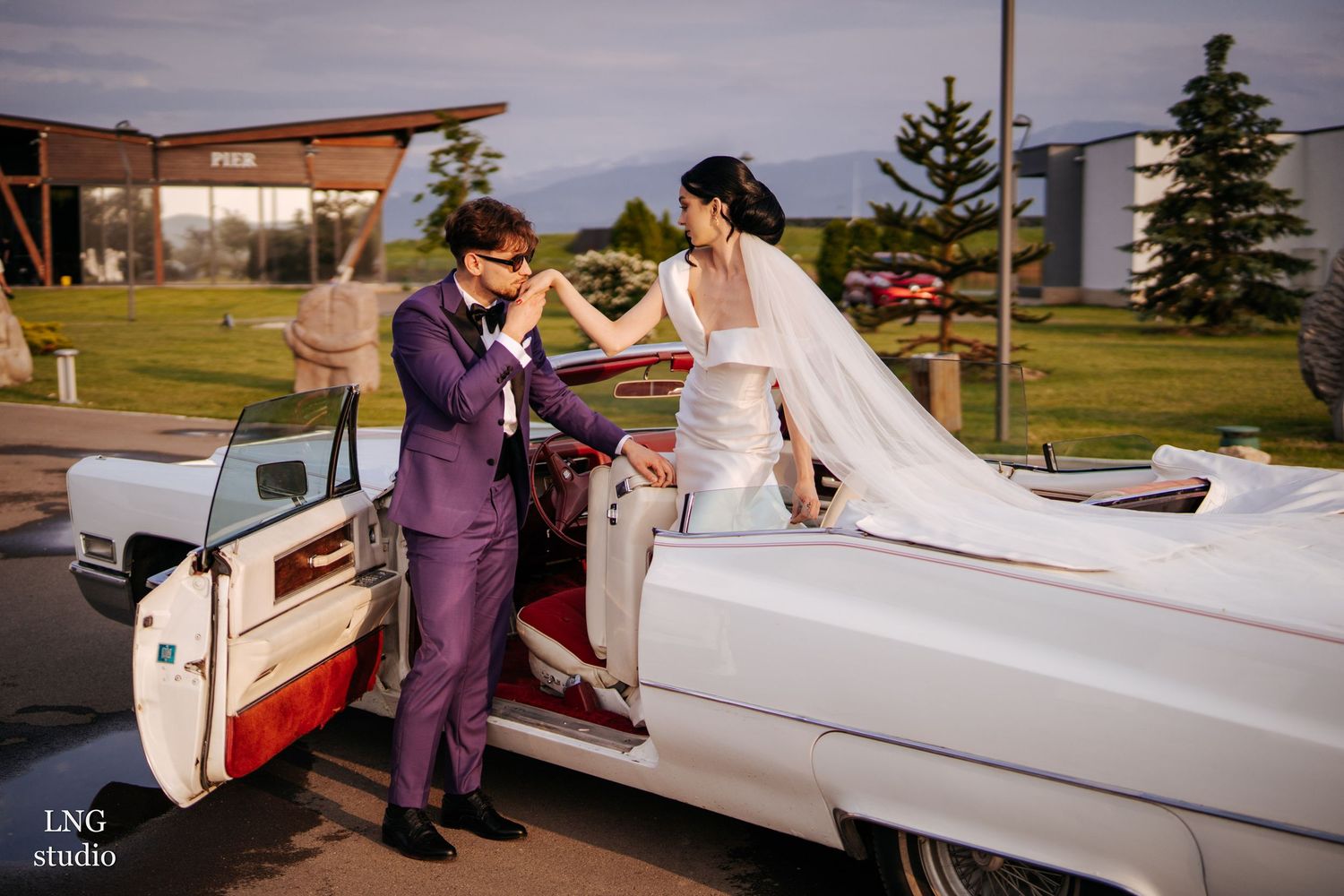 A wedding party wearing purple poses by a white classic car for formal photos.
