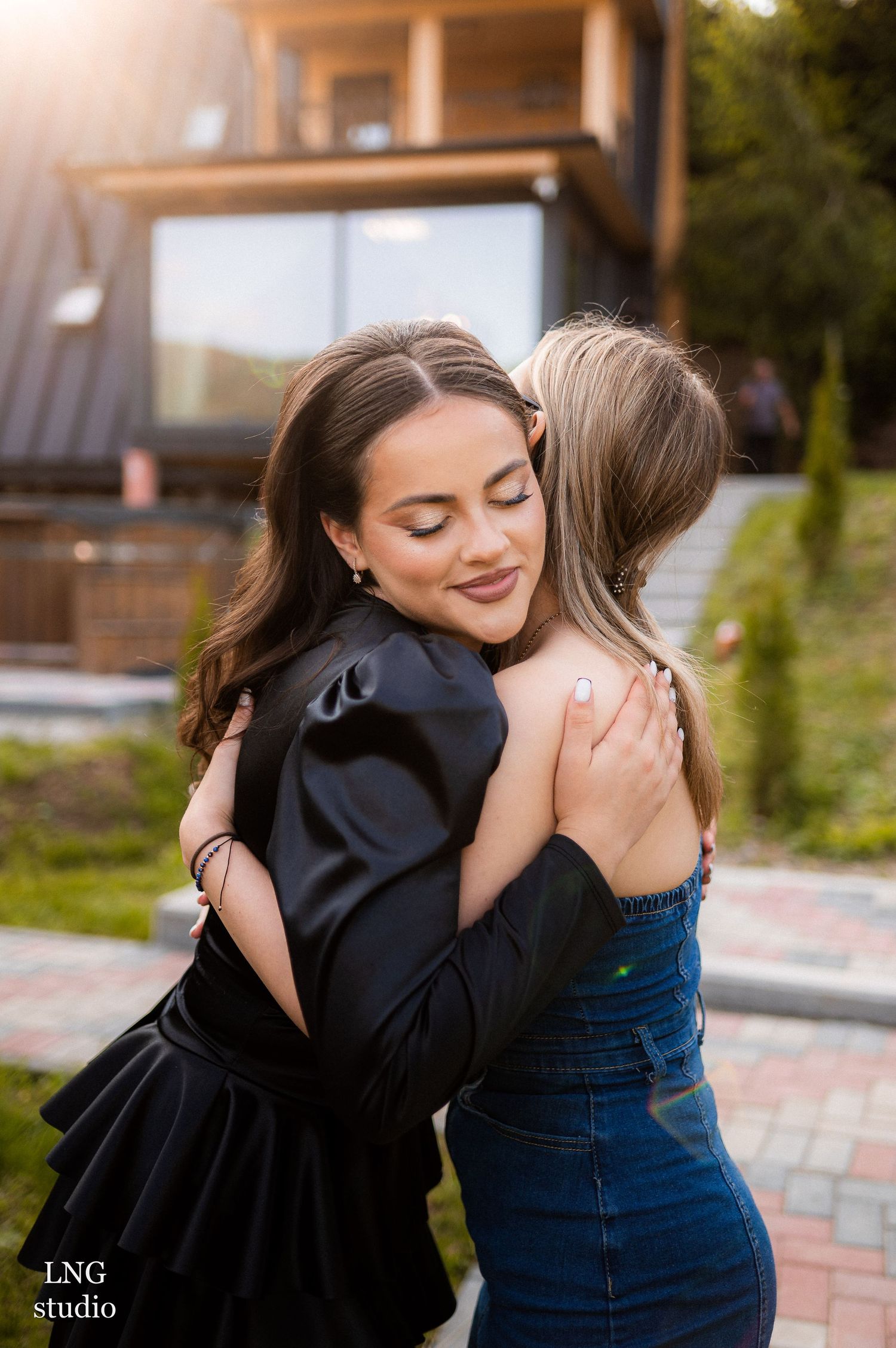 Two people share an emotional embrace wearing contrasting black and blue outfits in an outdoor setting at dusk.
