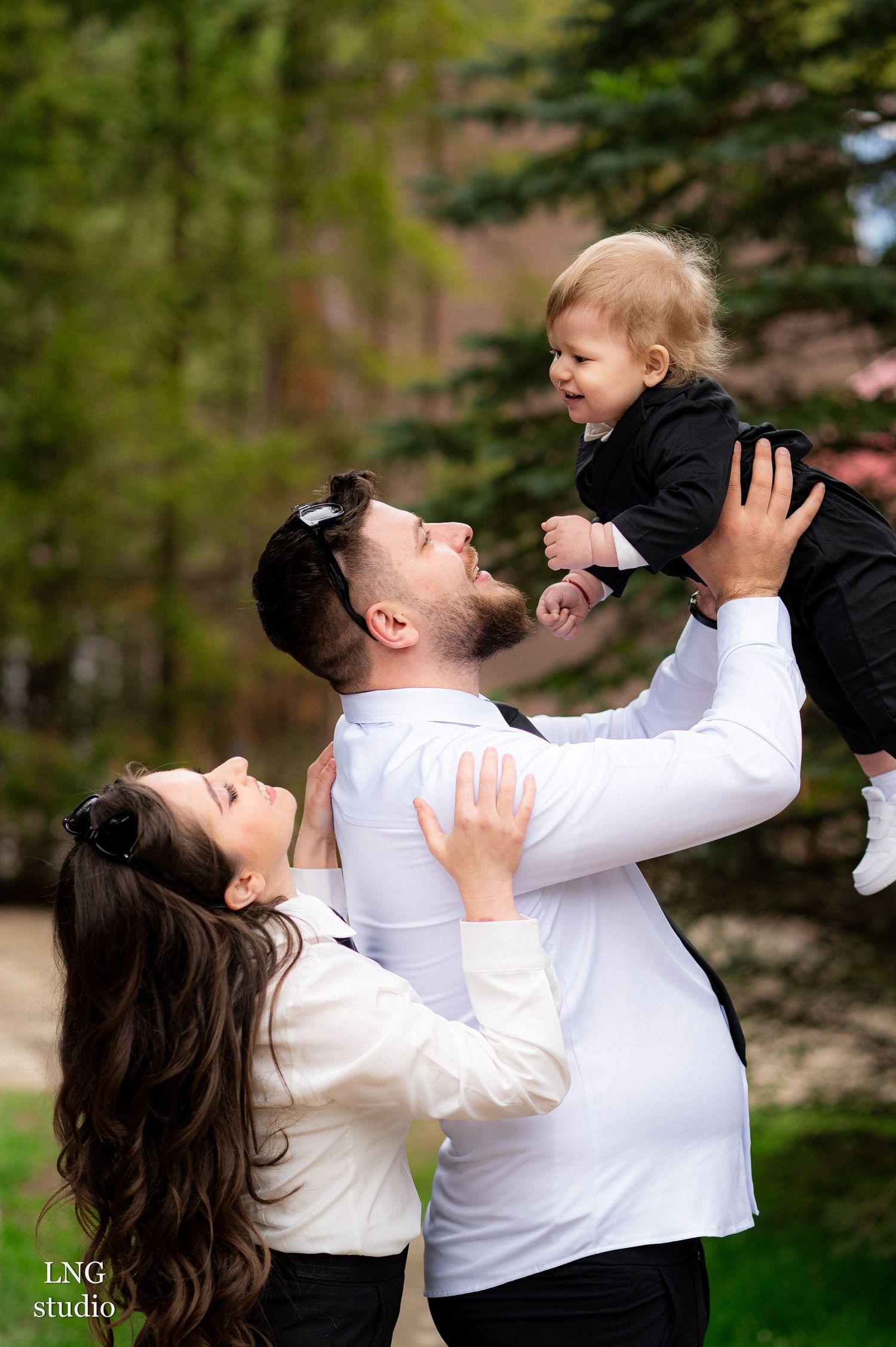 Joyful family moment captured outdoors with playful lifting pose against evergreen trees.