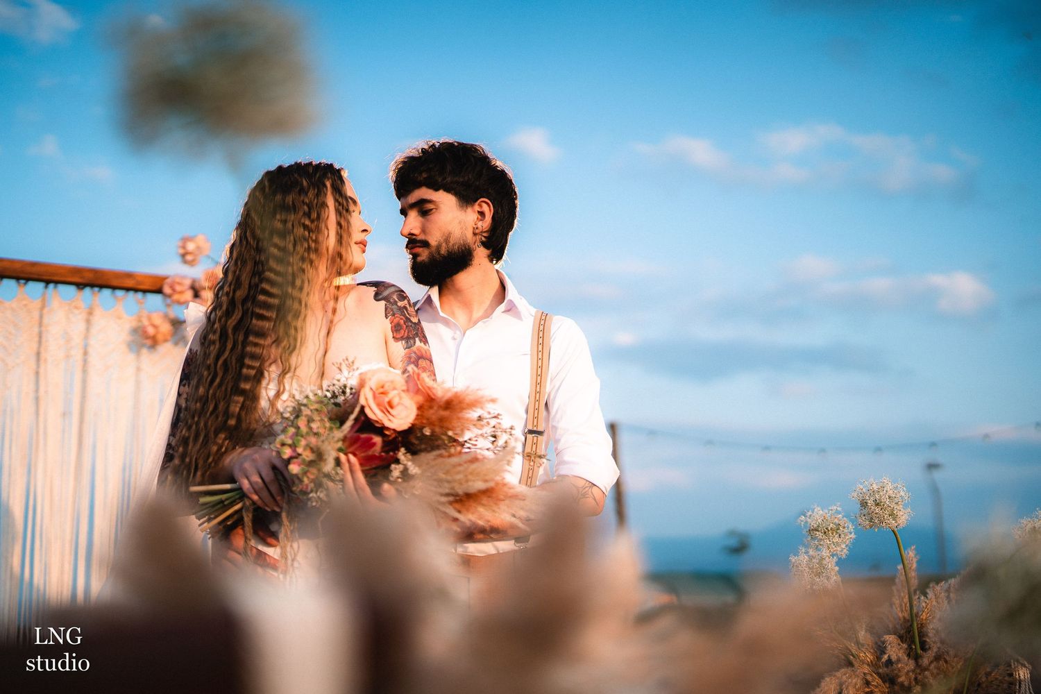 A couple embraces in scenic natural lighting while surrounded by dried flowers and sunlit skies.