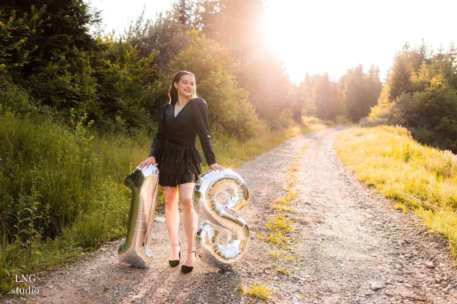 A dirt path through sunlit forest with silver number 18 balloons and a person standing at golden hour.
